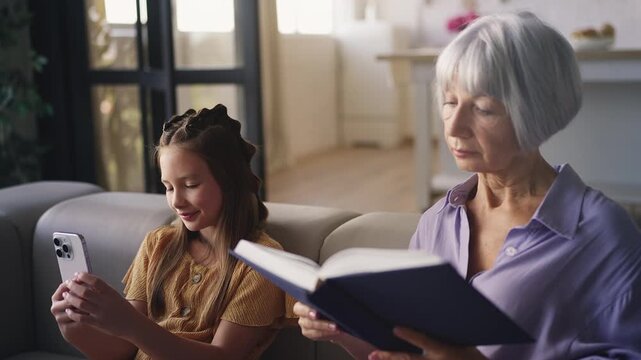 Smiling girl playing on smartphone while her granny reads a book at home