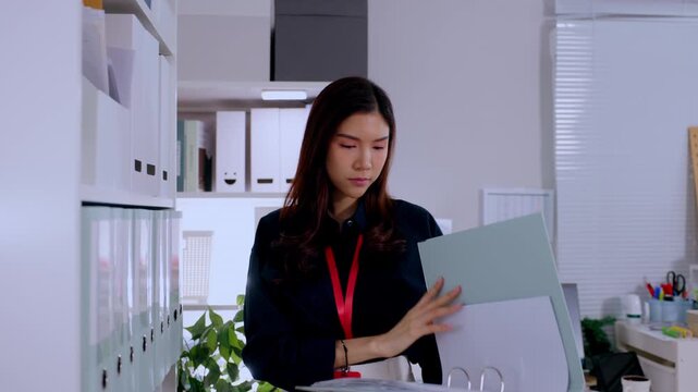Serious young adult asian woman holding open file folder searching corporate office archive shelf while professional female pulls paperwork binder retrieving important document during busy workday
