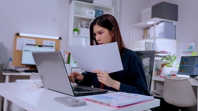Focused businesswoman sitting at modern office desk reading paper document typing on laptop computer feeling stressed about upcoming corporate project deadline managing career report seriously