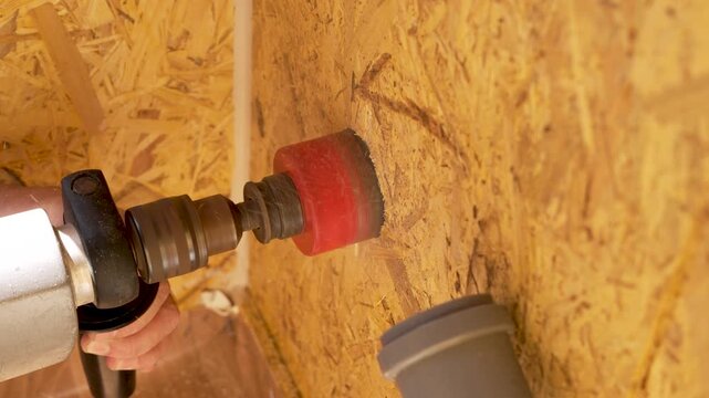 A power drill cuts a circular opening into an OSB wall panel during renovation work. The clip shows close-up woodworking, hole saw action, and pipe installation preparation.