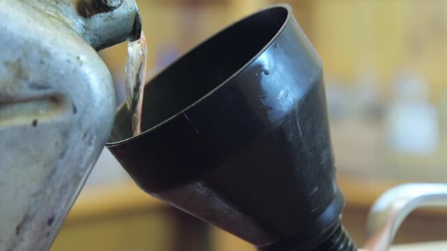 Pouring liquid from a metal container into a black funnel in a workshop setting during daytime