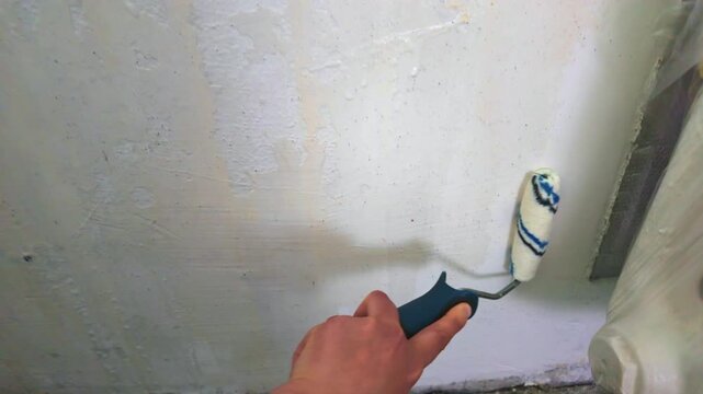 Person priming interior wall with roller near radiator and baseboard