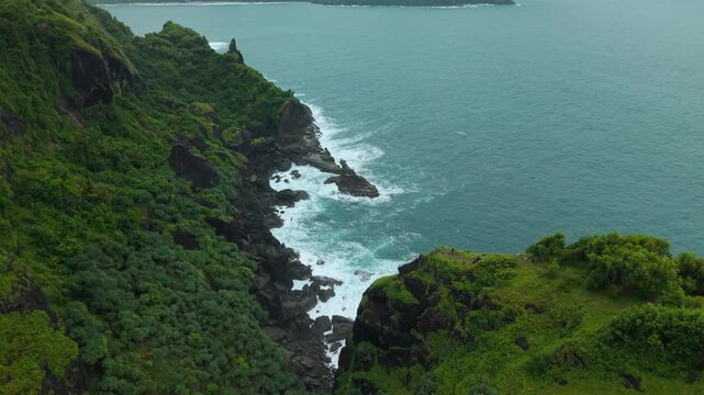 Aerial view of Bukit Pengilon featuring lush green cliffs and waves crashing against the rocky coastline in Kecamatan Tepus, Daerah Istimewa Yogya, Indonesia.