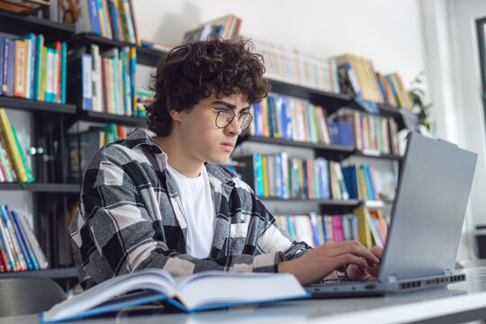 Smart curly haired teenage boy with glasses using a laptop while studying in a library, engaged in online learning, STEM coding education, and academic exam preparation.