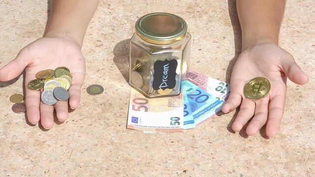Close-up conceptual video of a child learning financial literacy and saving money for a dream. In the frame, the child holds coins in one hand and a symbolic bitcoin token in the other