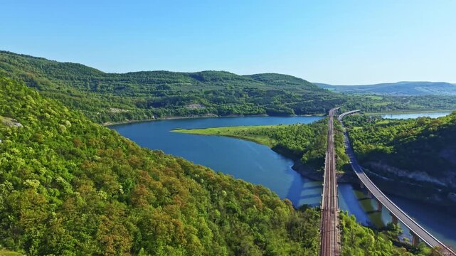 A river flows through green hills with trees. A railway track runs parallel to the river. The scene is bright and sunny with clear blue sky.