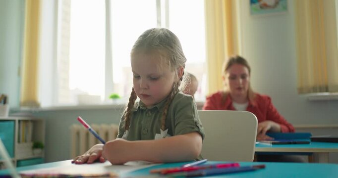 Young schoolgirl with braided hair concentrating on drawing at a classroom desk, with pencils, worksheets, classmates, and teacher in soft daylight