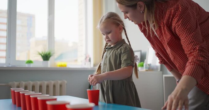 Young girl with braided hair and supportive woman concentrating on a colorful cup sorting activity at a table in a bright indoor classroom