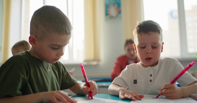Young children concentrating on drawing with colored pencils at classroom desks during a bright school art activity with classmates in background