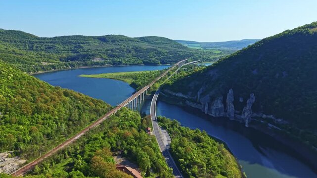 A long bridge spans across a wide river surrounded by green hills and trees. Sunlight shines on the water, creating reflections. The area shows rural landscape with clear skies.