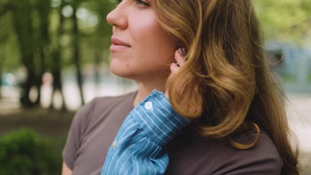 A close-up profile view of a thoughtful young woman fixing her wavy hair while standing outside in a green park. Female portrait, side perspective, natural beauty, reflection, casual lifestyle, calm m