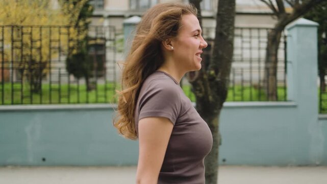 A happy young woman smiles while walking down a street in a profile view, holding her sunglasses. Female portrait, side perspective, city lifestyle, casual clothing, positive emotion, summer day.