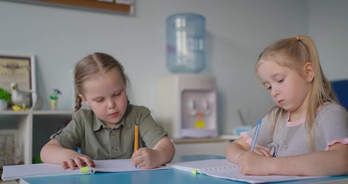 Two young girls concentrating on writing in notebooks at a classroom table, practicing schoolwork together in a calm early learning setting