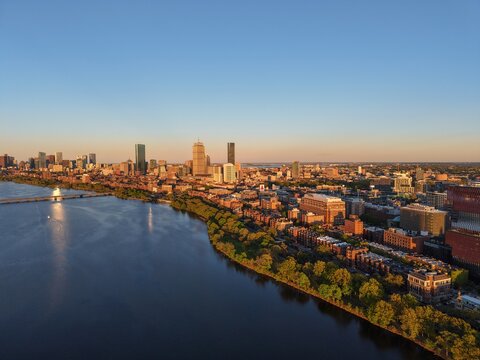 Golden hour aerial drone view of the Boston skyline, featuring the Prudential and John Hancock towers rising above the historic Back Bay neighborhood and the calm waters of the Charles River