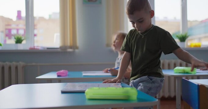 Young schoolboy concentrating at a classroom desk with colorful pencil case and notebook, surrounded by classmates in a bright learning space