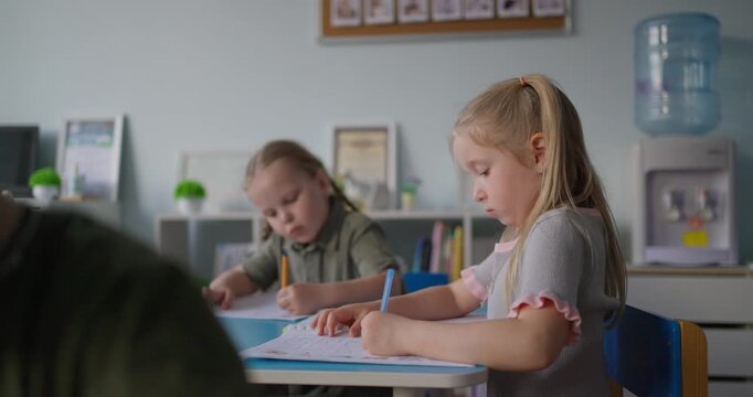 Young children concentrating on drawing and writing at classroom desks during a quiet preschool lesson, with a focused girl holding a pencil