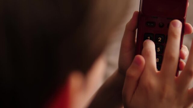 Over the shoulder view of a boy dialing a number on the keypad of a red retro telephone. The child presses the buttons of the vintage device while holding the receiver in his hands.