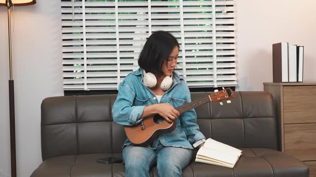 Young Asian woman wearing white headphones, smiling while looking down at her ukulele on a leather sofa in a cozy living room, practicing music or learning a new hobby concept