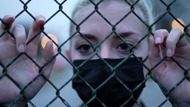Close-up video of young caucasian woman with pale blonde hair and black surgical mask, gripping a weathered chain-link fence, urban shadows bokeh background. Profound isolation and quiet contemplation