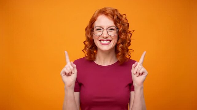 A cheerful young person with vibrant red hair, round glasses, and a dark crimson t-shirt smiles, hands raised, pointing up against a blurred orange studio background. Energetic celebration concept
