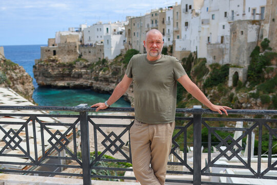 Mature man posing at scenic viewpoint in Polignano a Mare, Italy with historic cliffside town and Mediterranean coast background.