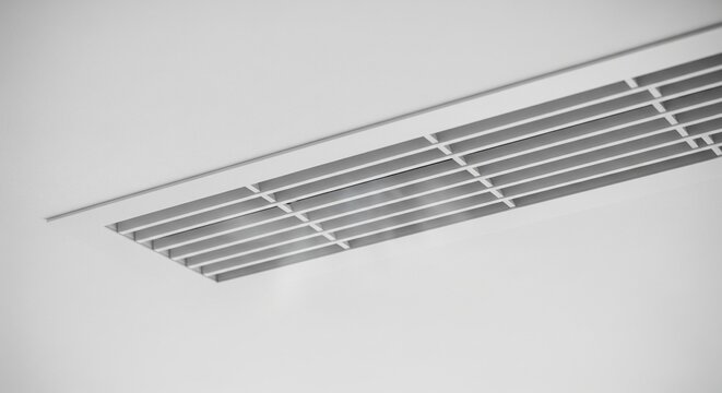 Black and white minimalist close-up view of a long linear slot air diffuser and ventilation vent on a plain white ceiling.
