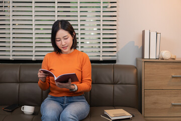 Fototapeta premium Happy young Asian woman sitting cross-legged on a leather sofa and smiling while reading a book in a cozy modern living room, educational or leisure hobby concept