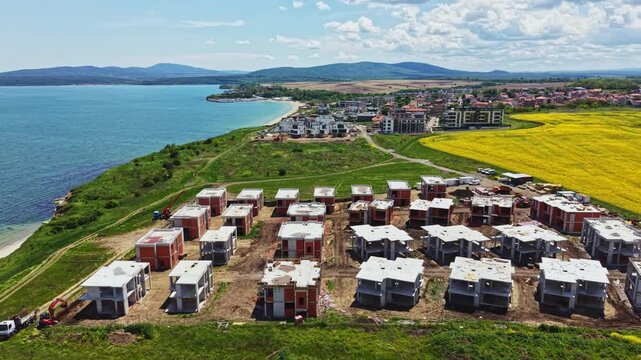 Builders work on new housing units by the coast in summer. The area is near a body of water, and a town is visible in the background. Fields are green and blooming.