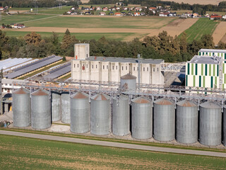 Drone view shows silos and farm buildings surrounded by fields and homes during daytime in a countryside location. © 24K-Production