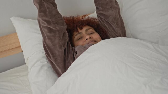 Woman with eyes closed stretching face and hands behind head in bed studio under white duvet and pillow; serenity.