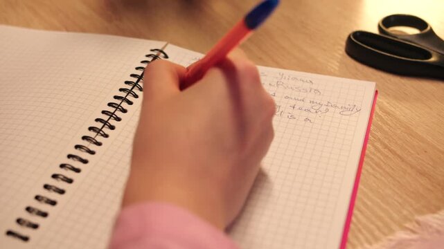 Closeup of female hand that writes personal diary entry with grid paper and spiral binding on table. Private reflection of woman captured in handwriting with orange pen in cozy evening scene