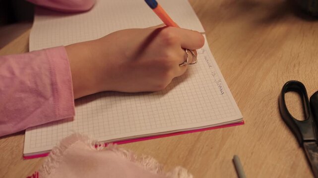Female hand writes in spiral notebook, cozy desk scene with warm lamp light, scissors and ring visible. Focused caucasian woman writes in grid paper, thoughtful journaling and study atmosphere