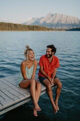 Fototapeta premium Couple sitting laughing on wooden dock by lake with mountain backdrop