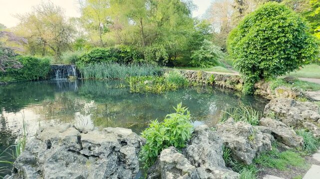 Weathered, rugged stones frame the calm water of an idyllic garden pond in the morning sun. A small waterfall cascades gently in the distance, bringing a refreshing feeling of peaceful solace.