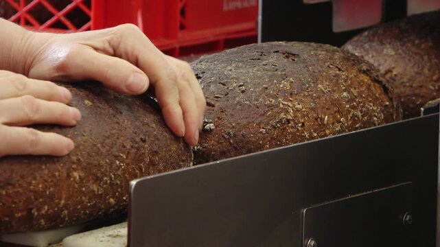 A bare hand steadies a large crusty loaf of dark bread on bakery slicing equipment, with red crates blurred in the background.