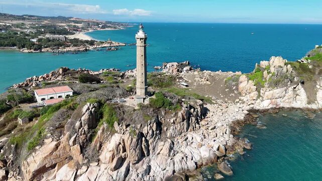 Aerial cinematic view of Ke Ga Lighthouse and its rocky island surrounded by emerald sea with panoramic coastal landscape in Binh Thuan, Vietnam.