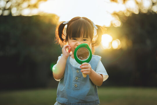 Cute little Asian girl holding magnifying glass at public park during sunset happy toddler exploring nature funny child looking through toy lens outdoor early education and curious discovery concept