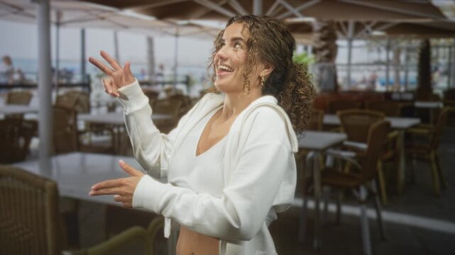 Young woman with curly hair smiles and gestures with hands, showing navel piercing in an outdoor seaside cafe by the boardwalk; joyful carefree.