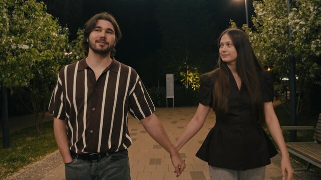 Couple takes quiet night walk in park beneath blossoms and glowing lights. Gentle movement and soft atmosphere suggest romantic leisure.