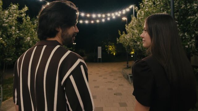 Young couple walks hand in hand through park at night before sharing a kiss. Romantic outdoor stroll creates calm intimate atmosphere.