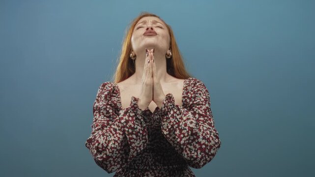 Redhead woman with clasped palms and tilted head, eyes closed, lips pursed while praying in a studio; hopeful devotion.
