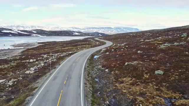 Aerial view of a winding asphalt road through the rugged plateau of Hardangervidda with snow patches and rocky hills Ullensvang, Vestland, Norway.