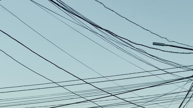 A complex web of black electric cables crisscrosses over a pale, clear sky above an urban road. The chaotic tangle of wires feels overwhelming, messy, and structurally stressful to look at.