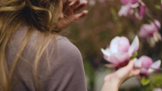 A close-up back view of a young woman with beautiful wavy blonde hair admiring blooming pink magnolia flowers in a garden. Female portrait, hair care, spring season, natural beauty, profile view
