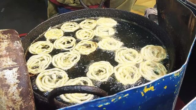 Golden jalebis frying in hot bubbling oil within a large rustic metal pan during cooking