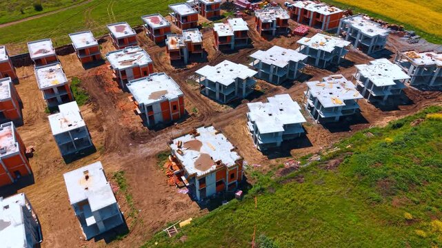 Aerial view of modern residential cottage community under construction. High angle view of multiple concrete and brick houses being built next to a green field.