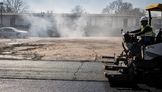 Medium shot of worker operating asphalt paver machine resurfacing motel parking lot with steam and dust in background bright daylight highlighting fresh pavement details.