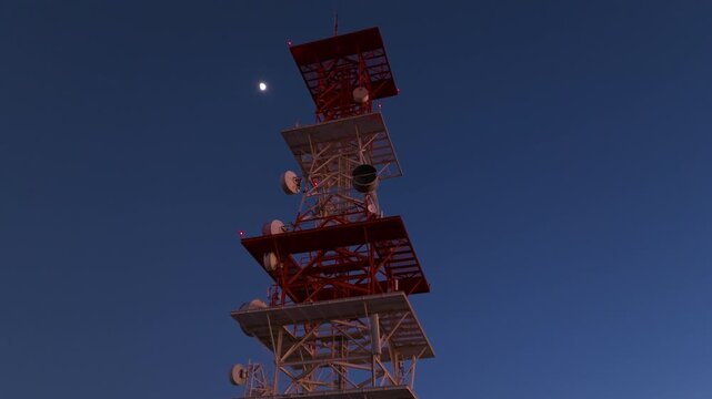 Tall communications tower with multiple satellite dishes and microwave antennas stands against deep blue dusk sky. Red aviation warning lights blink on metal structure with moon visible nearby.