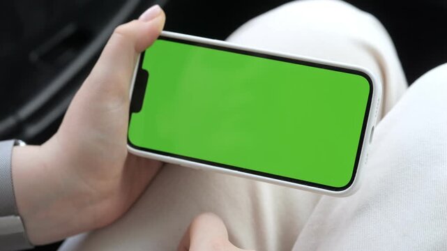 Hand holds phone with green screen. Person sits in car. Wears light-colored pants. Wears gray smartwatch. Phone frame silver. Background blurred