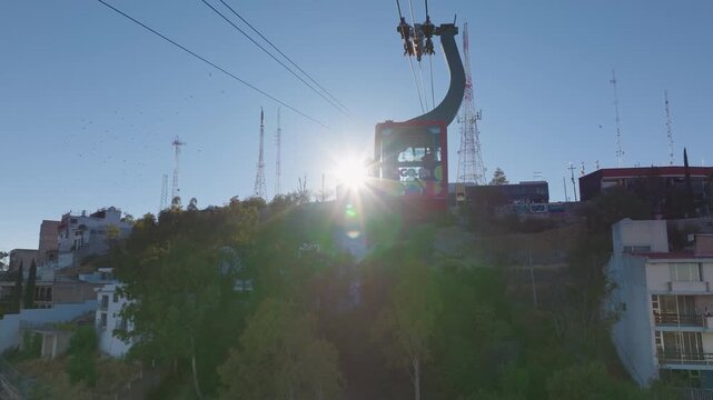 Red aerial tramway gondola travels uphill past telecommunication towers during golden hour. Bright sun creates dramatic lens flare. Urban hillside with buildings and graffiti walls visible below.